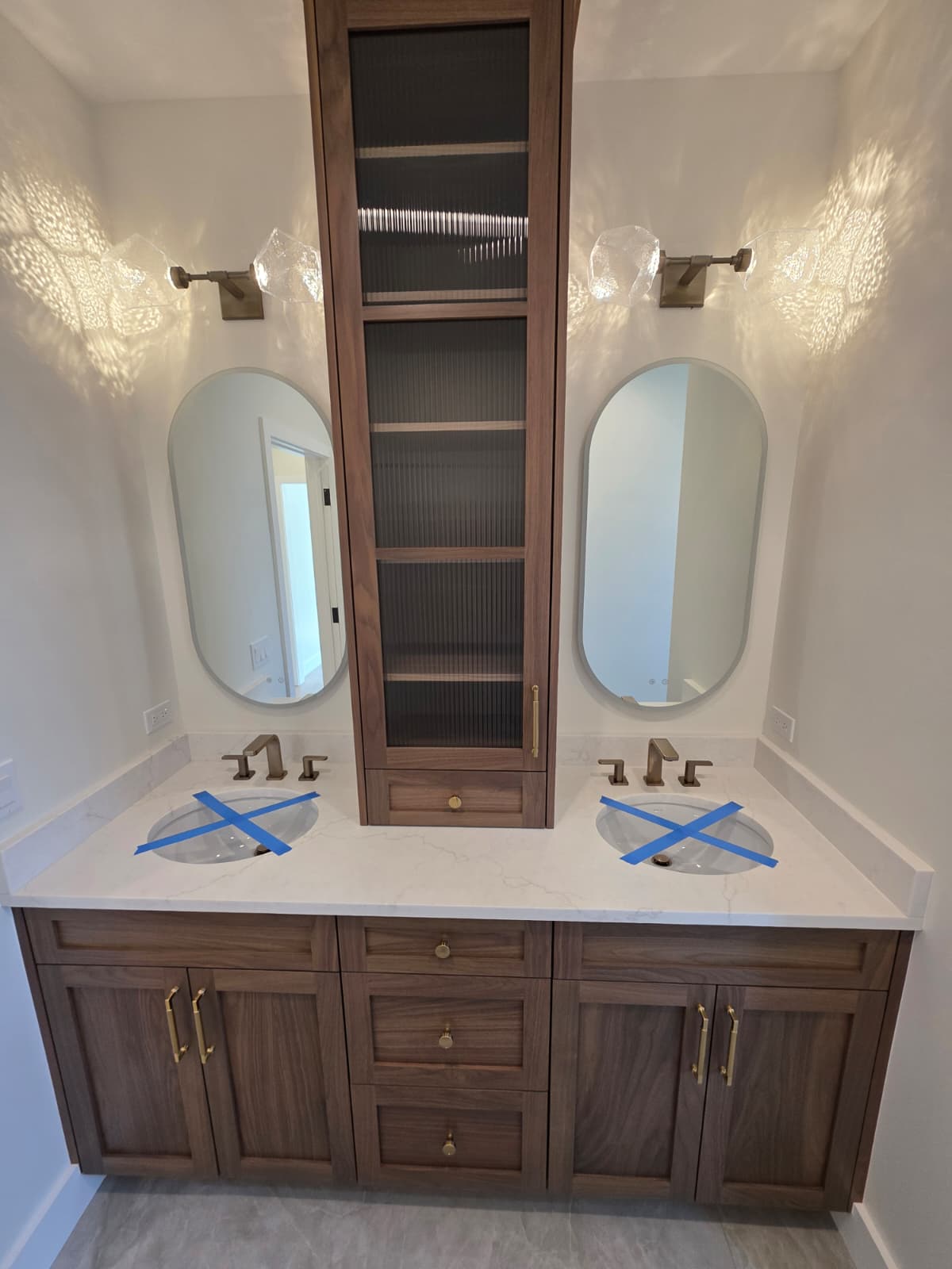 Walnut double vanity with oval mirrors, fluted glass storage tower and brass fixtures